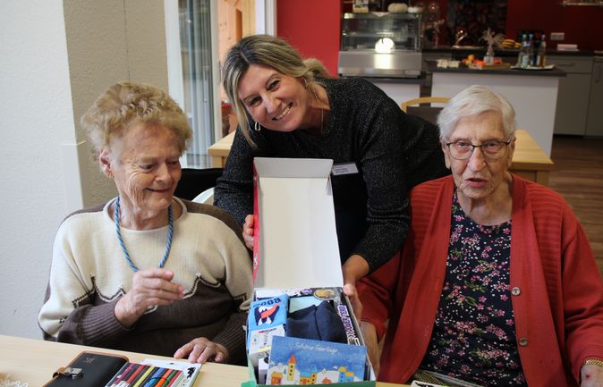 eine lächelnde Frau reicht zwei Seniorinnen einen offenen Schuhkarton mit Weihnachtsgeschenken