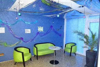 Wartebereich mit drei grünen Sesseln, kleinem Tisch, blauer Wanddekoration und Topfpflanze
