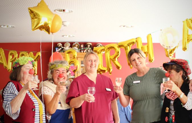 Hausclowns und Mitarbeitende stoßen vor Goldballons zum Geburtstag an