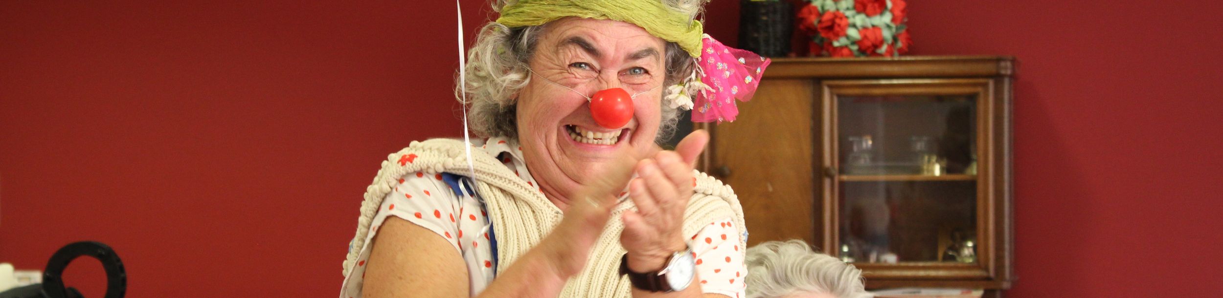 Frau im Clownskostüm mit roter Nase klatscht, Seniorin sitzt dahinter