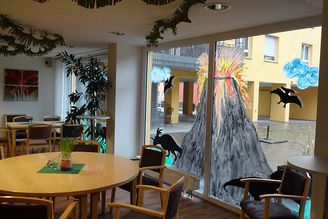 Aufenthaltsraum mit Fensterbemalung eines ausbrechenden Vulkans und Dinosaurier-Silhouetten