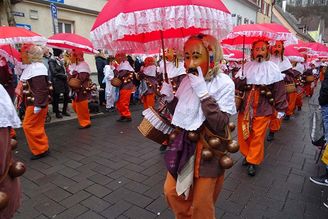 Maskierte Umzugsteilnehmer mit roten Schirmen und Glockengürteln