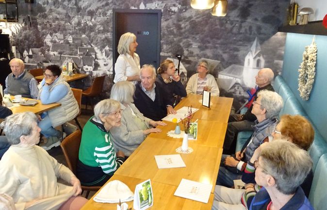 Seniorengruppe bei Tagespflege-Ausflug im Café mit Dorfwandbild