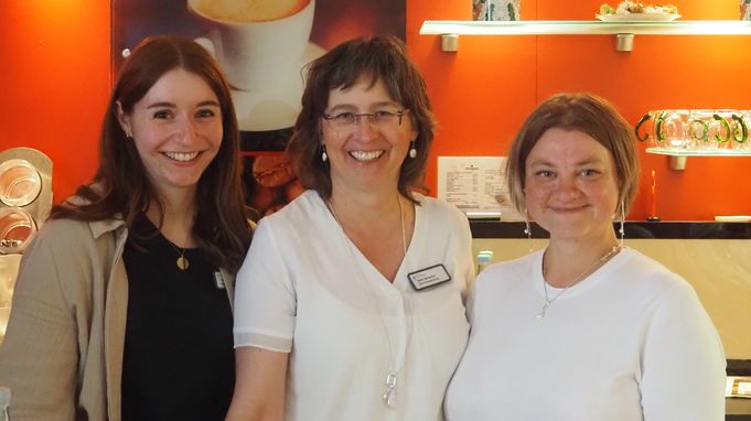 Drei lächelnde Frauen vor roter Wand mit Kaffeeplakat
