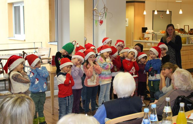 Kindergartenkinder mit Weihnachtsmützen stehen vor Senioren im Gemeinschaftsraum
