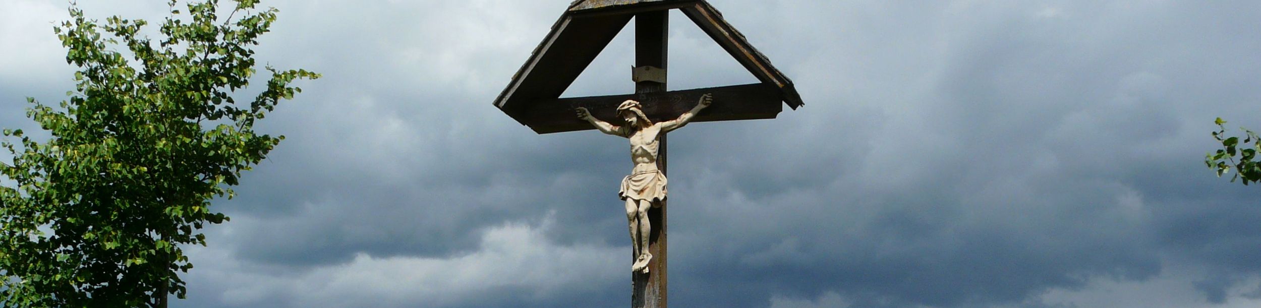 Holzkreuz mit Korpus vor Getreidefeld und dunklen Wolken